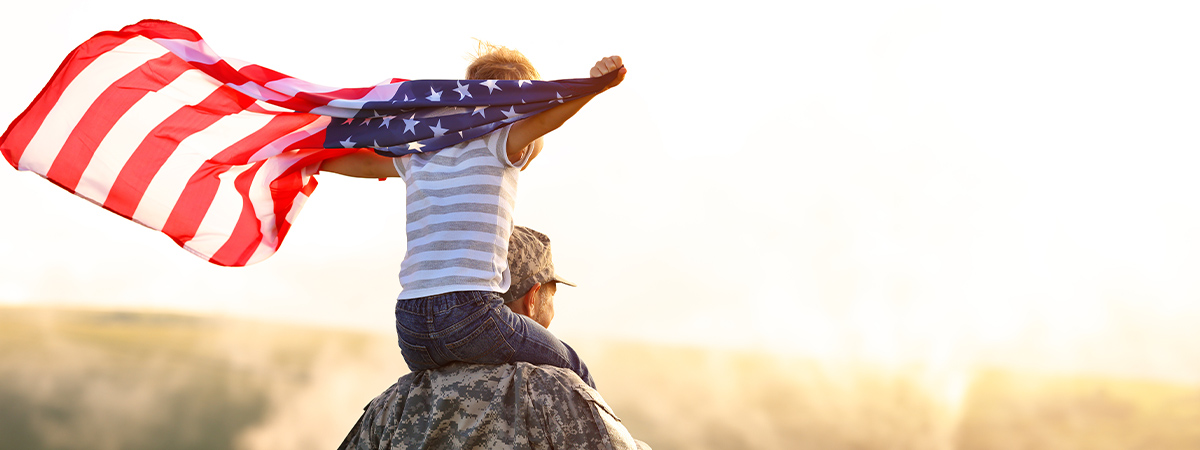 photo of a soldier and his kid on his shoulders holding a flowy American flag at a wheat field by sunset